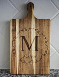 Wooden cutting board with engraved letter 'M' and 'Est 1998' on a tiled wall background