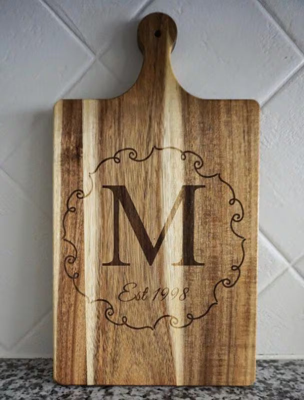 Wooden cutting board with engraved letter 'M' and 'Est 1998' on a tiled wall background