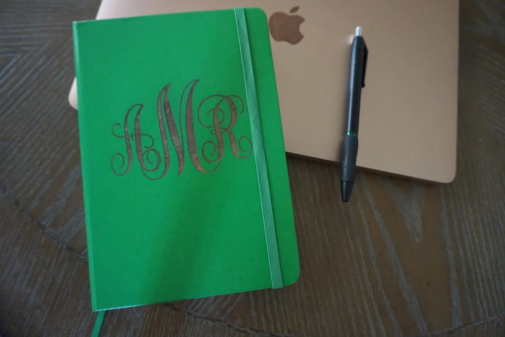 Green notebook with monogram initials on a wooden surface next to an Apple product.