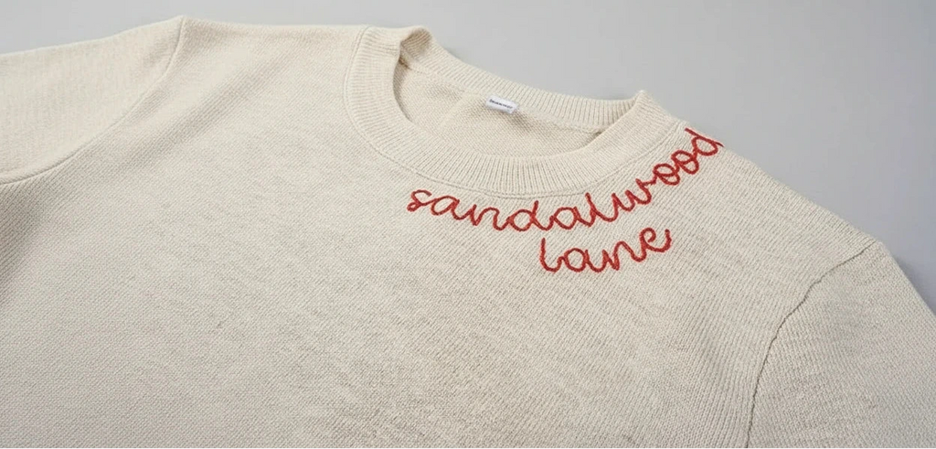 Beige sweater with red text 'sandalwood lane' on a wooden surface