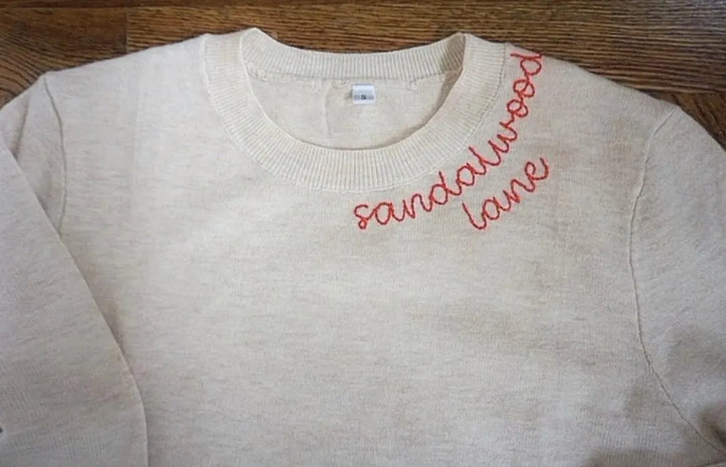 Beige sweatshirt with 'sandalwood lane' embroidered in red on a wooden surface