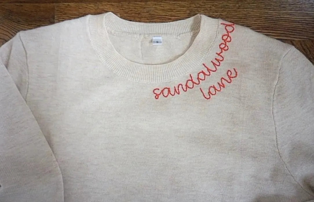 Beige sweatshirt with 'sandalwood lane' embroidered in red on a wooden surface