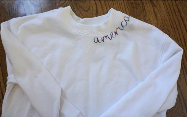 White sweatshirt with 'america' embroidered on it, laid flat on a wooden surface.